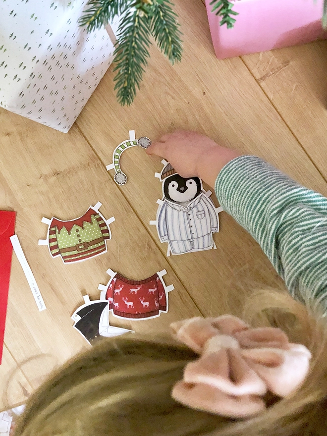 A young child is playing with a penguin paper doll on a wooden floor beside wrapped gifts.