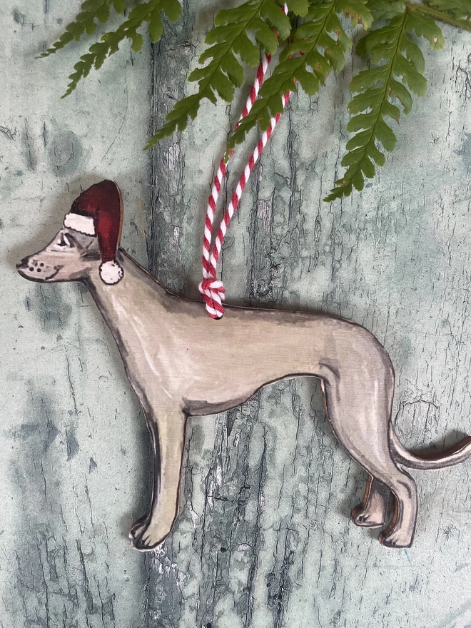 Whippet wooden Christmas decoration wearing a santa hat hung with red and white twine placed onto a red book about dogs