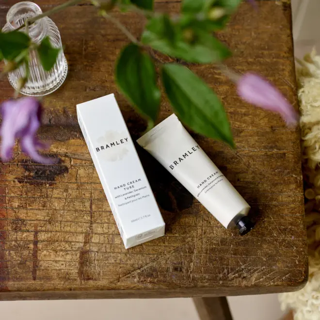 white tube of hand cream lying next to a white box of the same size, both reading 'bramley' and sitting on a wooden table, with a green plant in the foreground