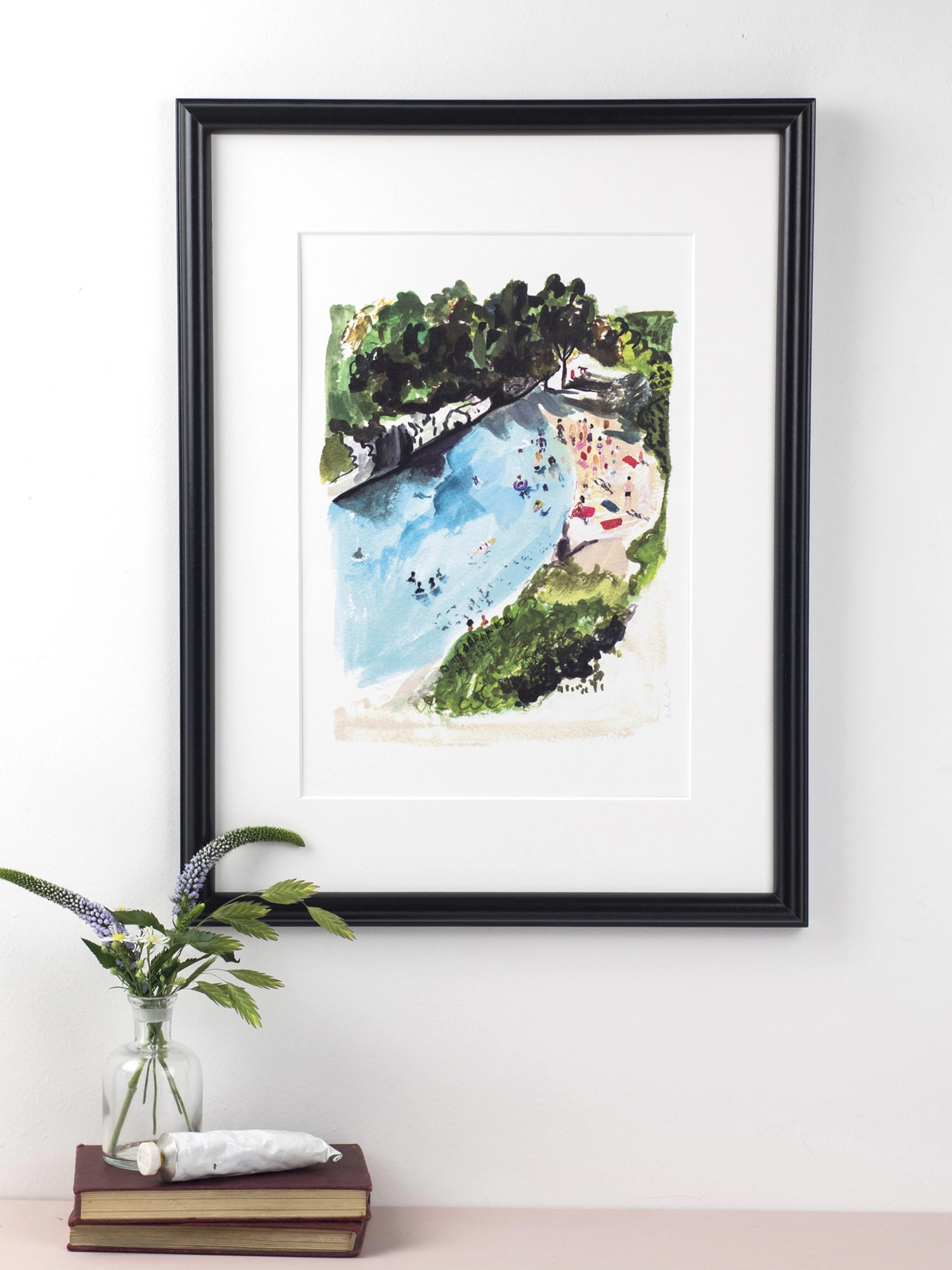 An art print featuring an observational drawing of a rocky beach in France. The small cove is full of people sunbathing and surrounded by trees. There are a few people in the water swimming.