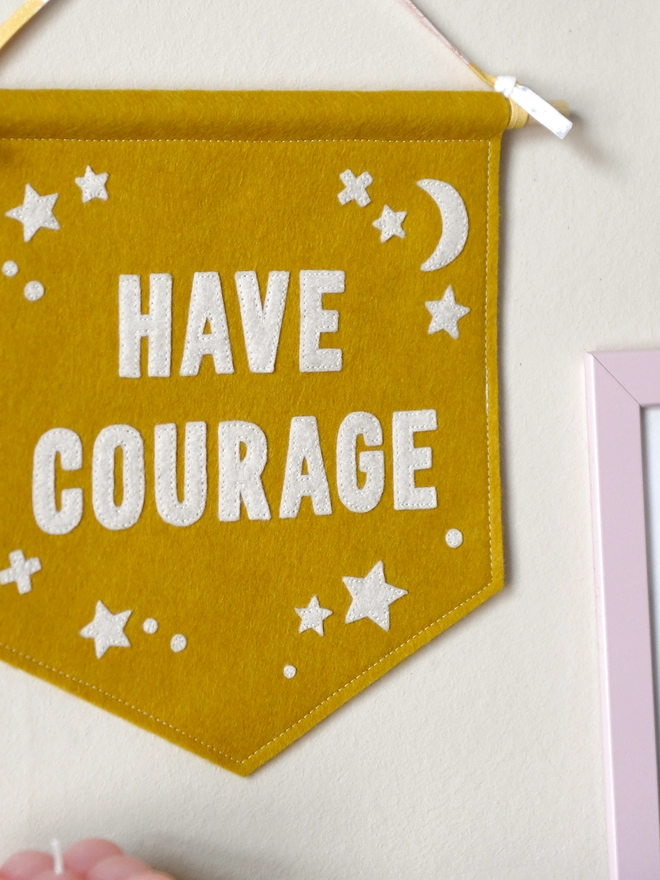A mustard yellow banner flag, with the words Have Courage in white, hangs on white wall by it’s ribbon attached to a wooden dowel at the top of the flag.