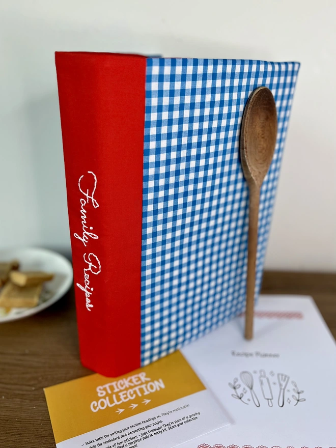 Blue gingham recipe binder with 'Family Recipes' embroidered on red spine, standing on wooden bench with wooden spoon propped against it and recipe template pages and 'Sticker Collection' envelope lying beside it. Plate of baking in background.
