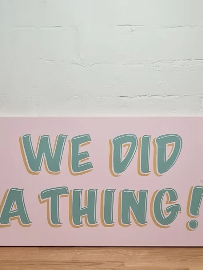 'We Did A Thing' Colourful Wedding Sign, a colourful wedding sign sitting on a wooden surface against a white brick backdrop. 