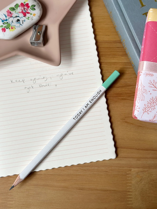 A white pencil, with a green end, and the words "Today I Am Enough" along the side lays on an open lined notebook on a wooden desk.