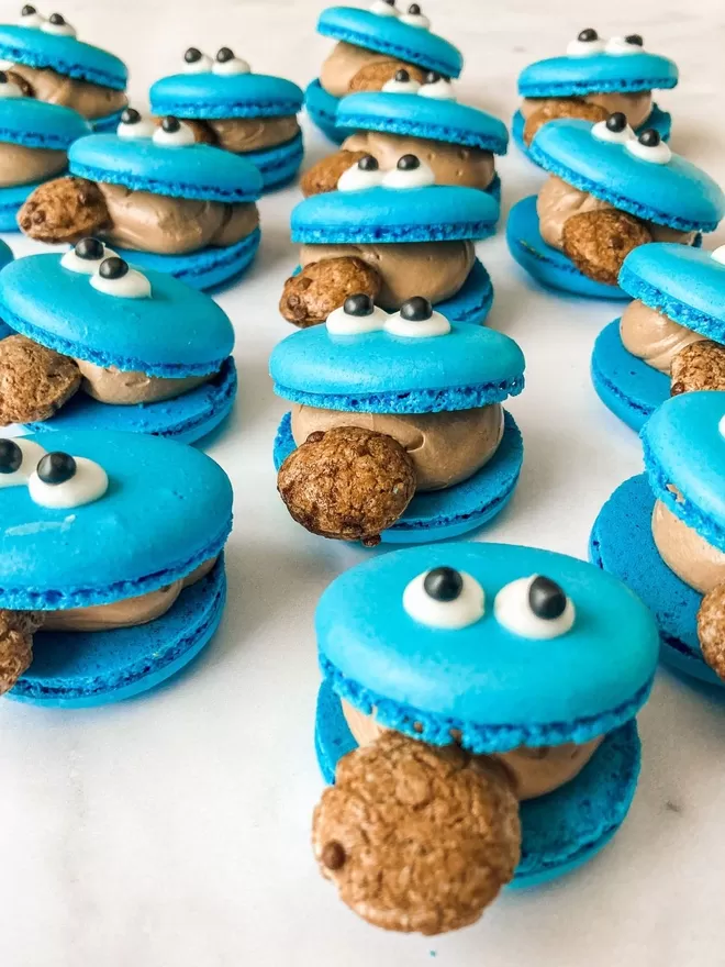 blue cookie monster macarons with googly eyes and a chocolate chip cookie in its mouth, lined up several rows