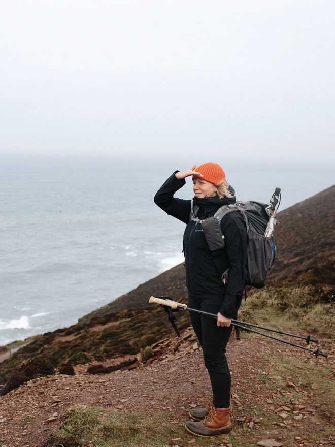 Woman wearing hiking gear and an orange hat, standing on hill by the sea 