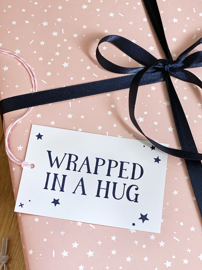 A gift wrapped in pink and white star wrapping paper has navy blue ribbon and a tag that reads "Wrapped In A Hug".