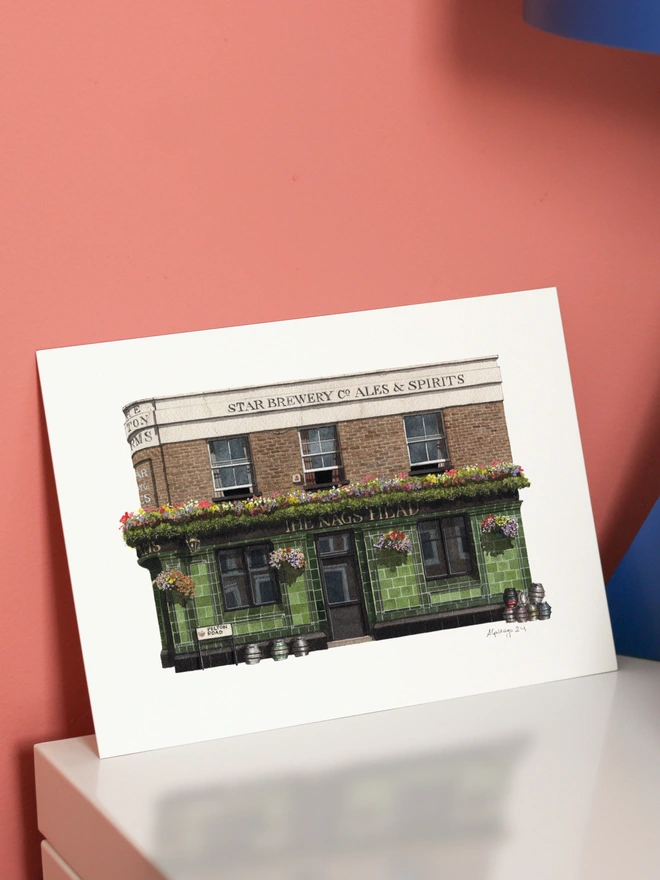 An architectural illustration of The Pelton Arms pub on a print, featuring green tiling and flower boxes, displayed on a white surface against a pink wall.