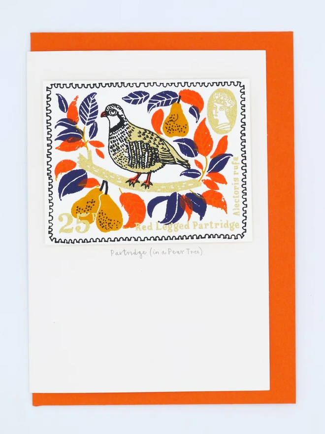 Partridge In A Pear Tree Christmas Card, a white card with a colourful bird design in the centre. It sits on top of an orange envelope on a plain surface. 