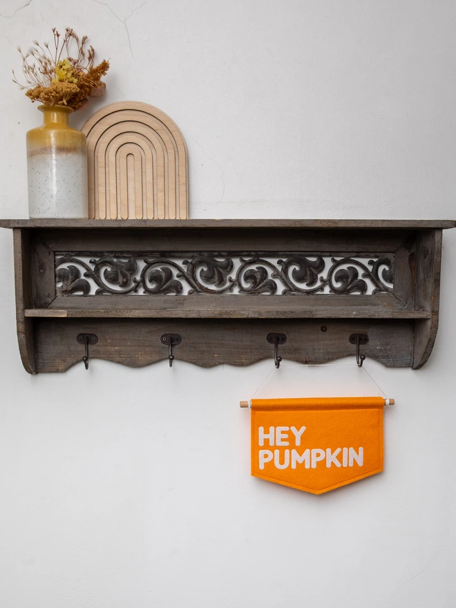 Orange, felt banner with the words "Hey Pumpkin" written on it in white glitter felt. The banner is hanging from a boho themed shelving unit with an autumnal flower arrangement and a wooden rainbow