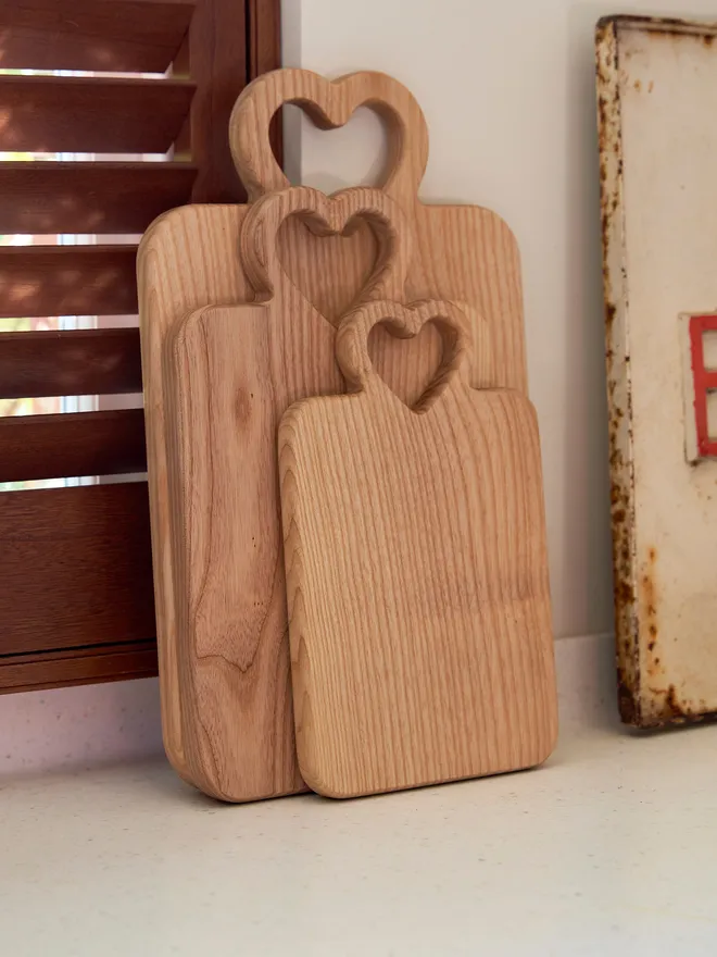 Set of 3 heart wooden serving boards leaning against a white tiled wall with brown shutters