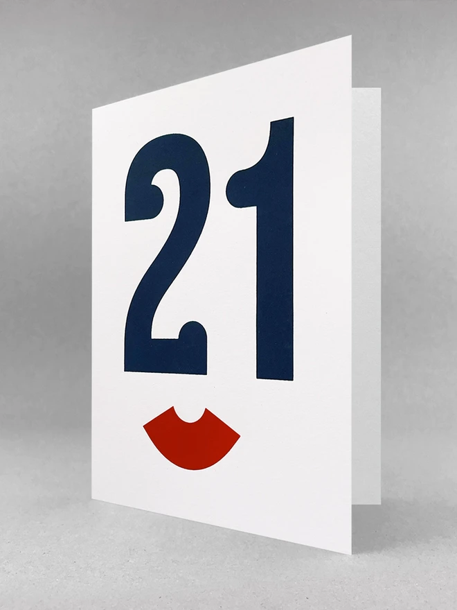The Number 21 screen printed in a dark blue serif typeface. The serifs make the number 21 look like the eyes and nose of a face from a slight angle. Below is a red bracket on its side. This looks like the lips of the face. The image has an Art Deco feel to it. The portrait card is stood slightly open, in a light grey studio set.