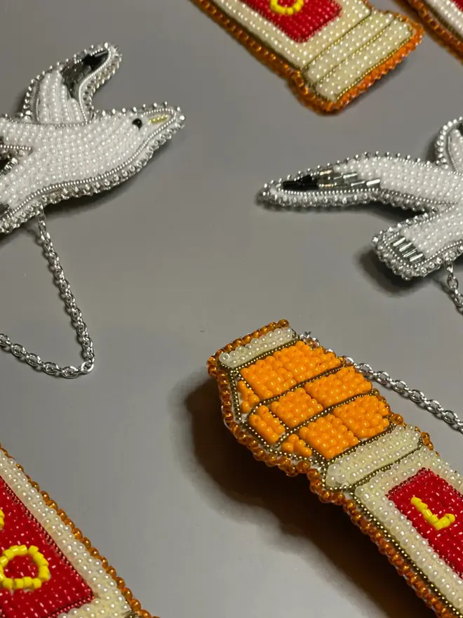 Beaded lido brooch with orange, red, and beige beads attached to a white beaded seagull by a silver chain, displayed on a grey table. 