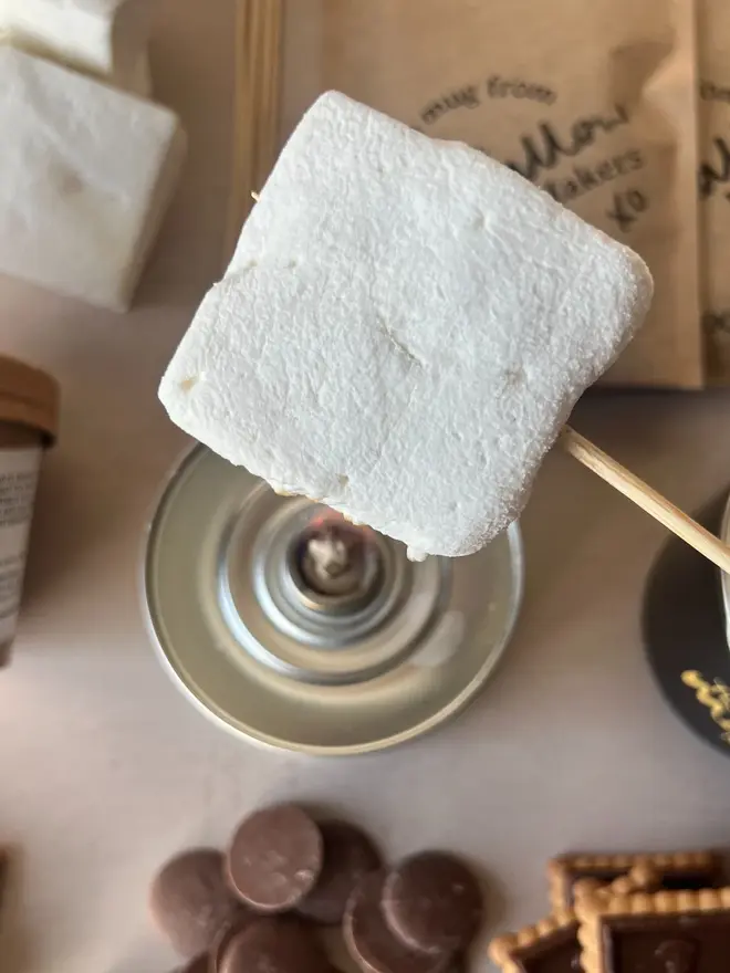 Luxury S'mores & Hot Chocolate Kit, a marshmallow skewered above the kit. This is laid out on a marble surface. 