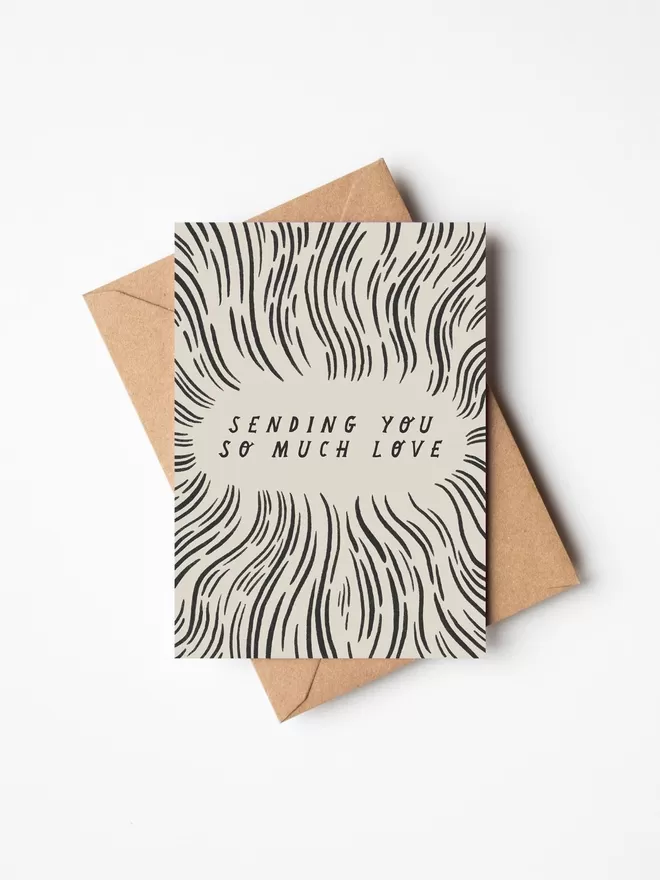 Black and white greeting card with illustration and the words sending you so much love on it with a brown envelope underneath it. 