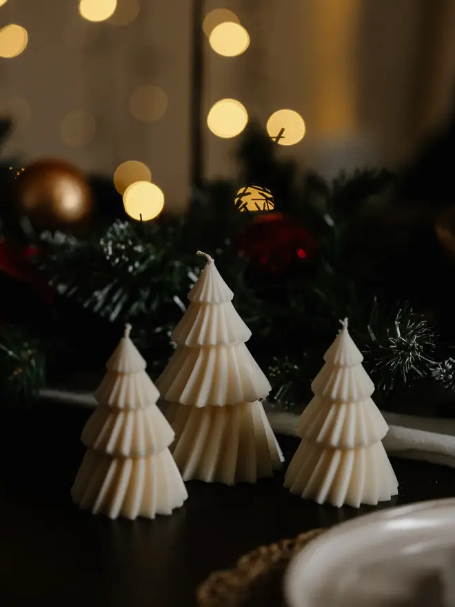 White Christmas Tree Warm Candles (Set Of 2). Three unlit cream tree candles in graduating heights on a dark table, with frosted pine garland, baubles and golden bokeh lights behind. The fluted design gives a soft, Scandi-inspired look to holiday tablescapes.