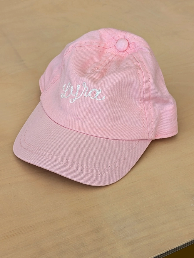 Baby Pink Toddler Baseball cap reading 'Lyra' in White embroidery, sitting on a wooden bench