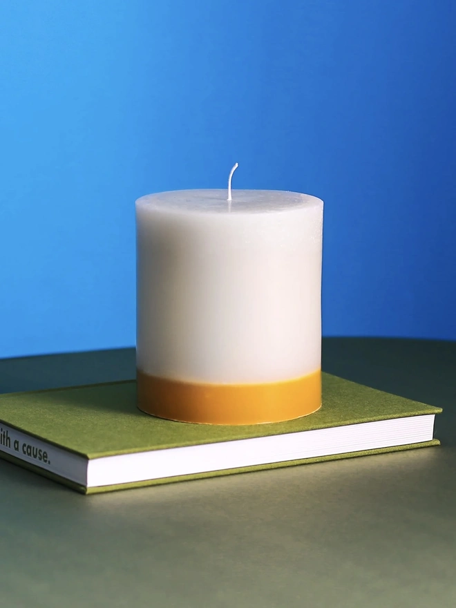 Jasmine, Green Tea & Mulberry Candle (Set Of 1 Or 3 Wicks). the jasmine, green tea and mulberry candle sits on a green book against a bright blue background, showing its single wick and cream wax with a golden yellow base. the two-tone colouring and textured, slightly uneven wax surface give the candle a rustic, hand-poured finish. the bold blue background highlights the candle's warm, honeyed tones.
