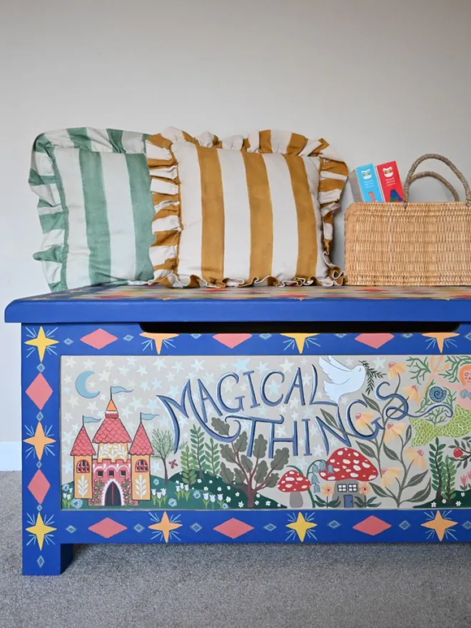 A colourful folk art wooden chest with a hinged lid, painted with stars and whimsical illustrations, including a castle and mushrooms, and the words "Magical Things" in the center. Two striped pillows and a woven basket sit on top.