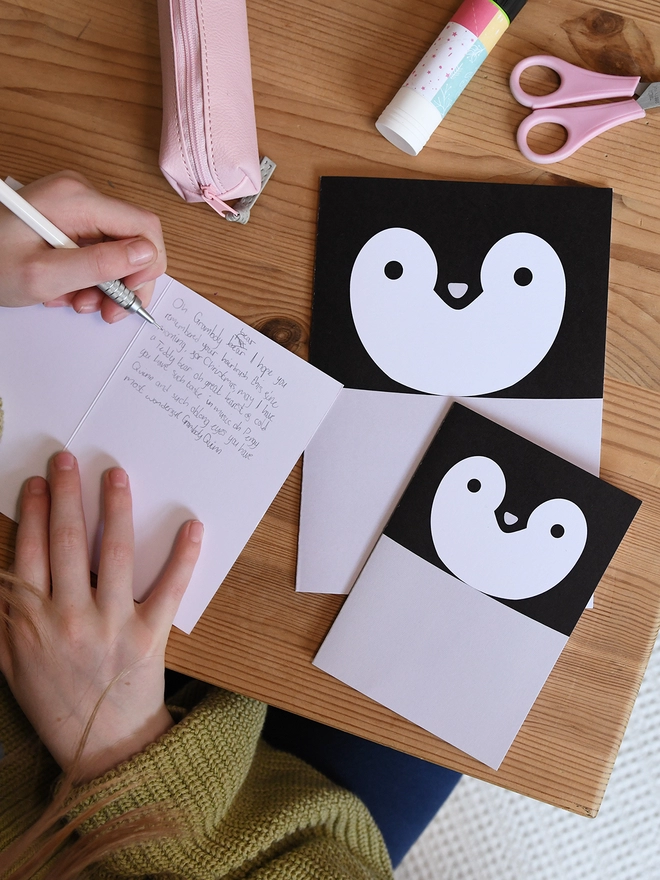 Several penguin greetings cards lay on a wooden table. A child is writing a message in one of the cards.