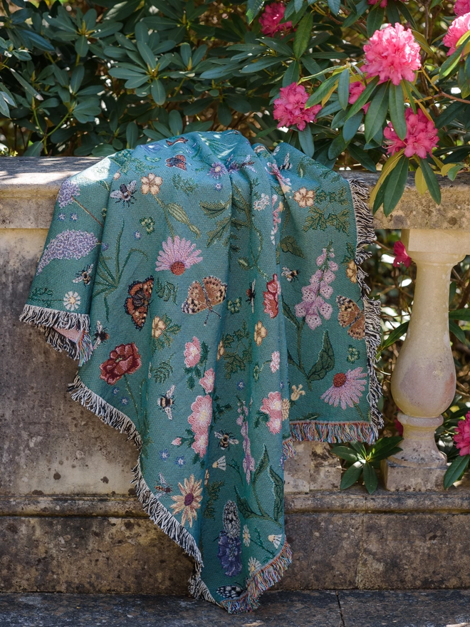 'Pollination Bloom' Recycled Cotton Blanket seen on a bannister with pink plants.