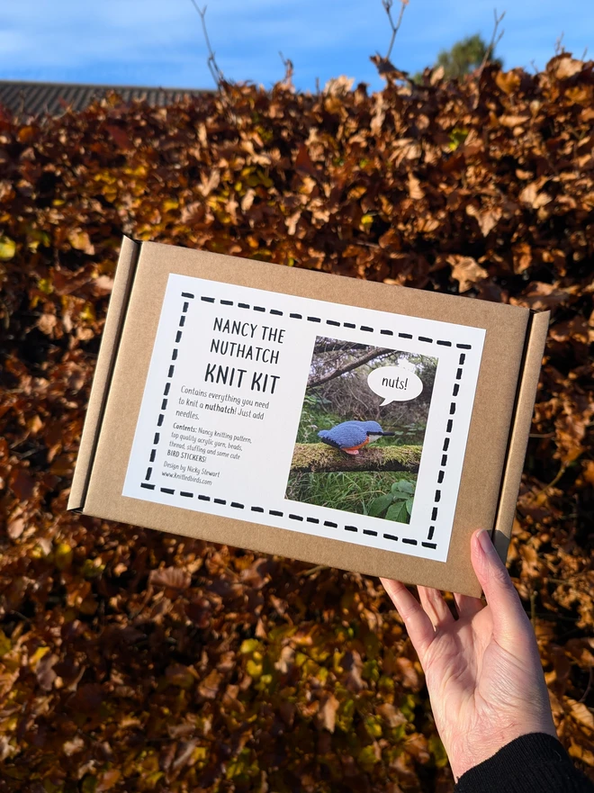 nuthatch bird knitting kit by Nicky Stewart. A knitting kit in a gift box being held up in front of a brown autumn beech hedge