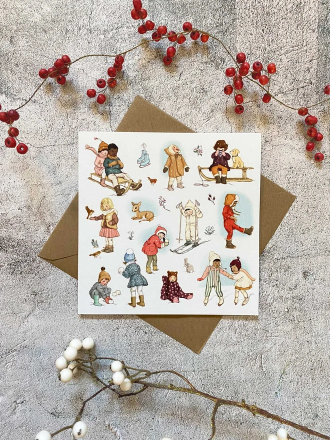 a Christmas greeting card featuring a vintage story book style illustration of many children playing in the snow. card is laid on a Kraft envelope