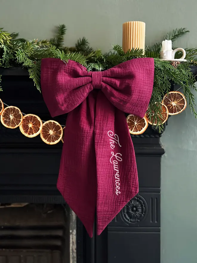 A handmade Christmas bow made from deep burgundy cotton with the words "The Laurences" on one tail is hung from a black mantlepiece that is decorated for Christmas with greenery, orange slice garland, and candles.