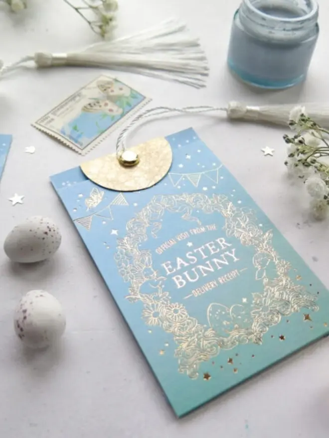 An angled view of the blue receipt pouch with its gold foil wreath of flowers, bunnies and eggs. White tassels and a butterfly postage stamp lie behind it.