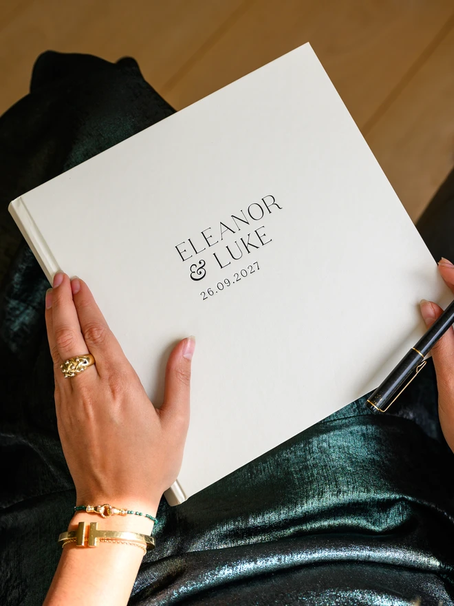 A pearl-coloured wedding guest book, personalised with printed lettering on the cover, rests on a woman's lap.