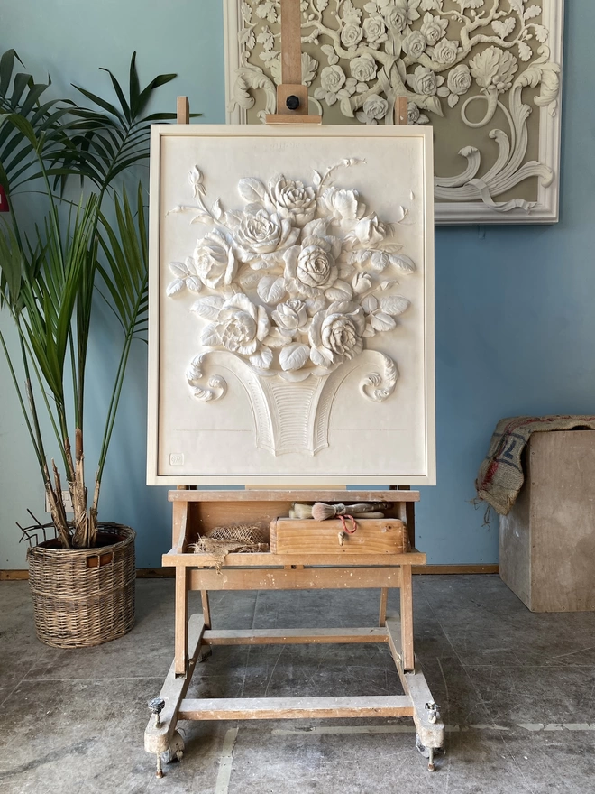 Plaster bas-relief flower sculpture on an easel with blue wall and potted plant behind