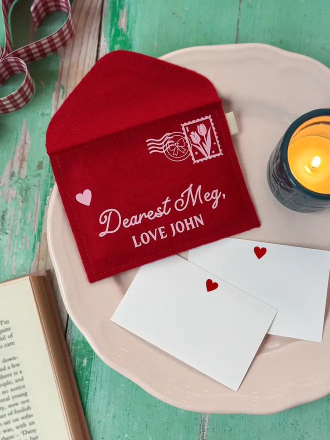 A handmade personalised red felt love letters envelope with the words 'Dearest Meg, love John' is displayed on a vintage-style plate with small heart cards, styled on a rustic green wooden table for Valentine’s Day.