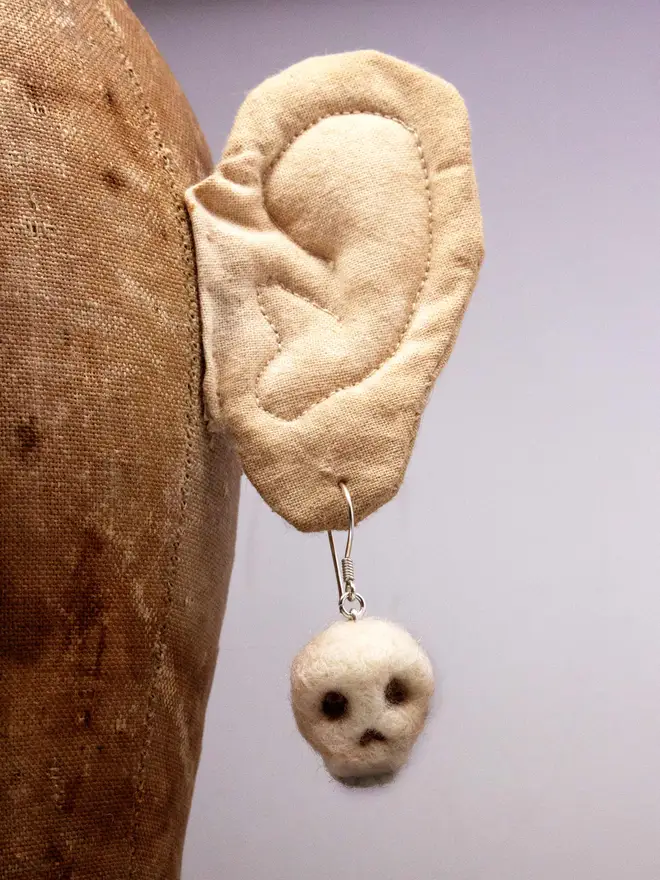 A needle-felted skull brooch dangling from a calico ear on a vintage hat display. Made by the artist Laura Cronin aka Bumble & Earwig