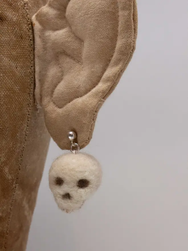 A needle felted fluffy skull earring on a fabric ear.  Made by the artist Laura Cronin aka Bumble & Earwig