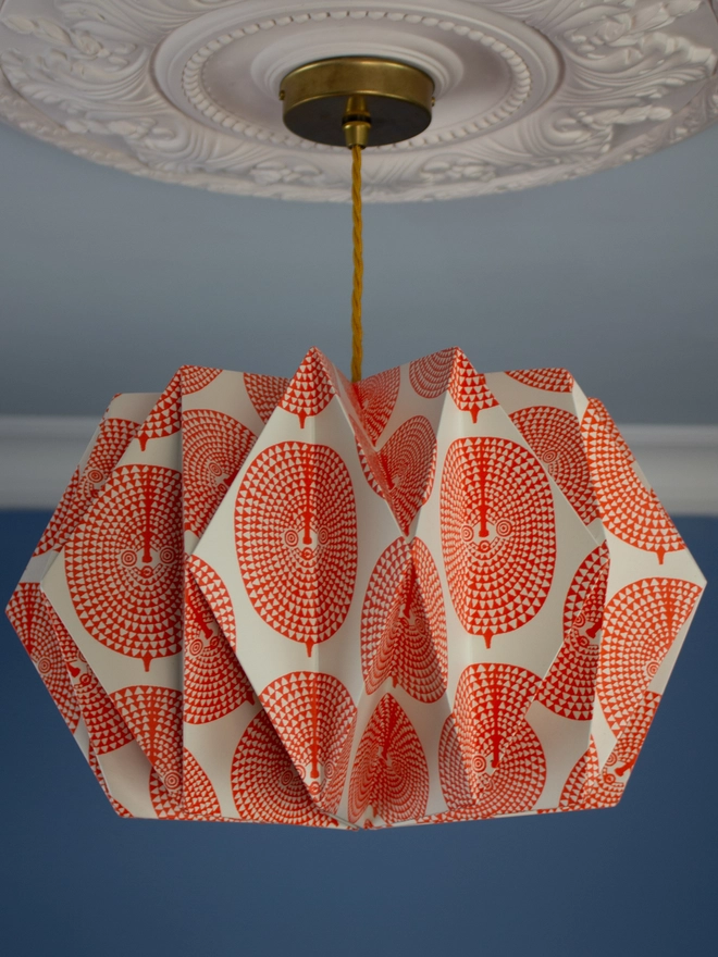 A vintage-inspired paper lampshade with a bold geometric pattern adds a touch of retro flair to a room with a blue wall and ornate ceiling. The lampshade is made from folded paper, creating a unique and eye-catching design.