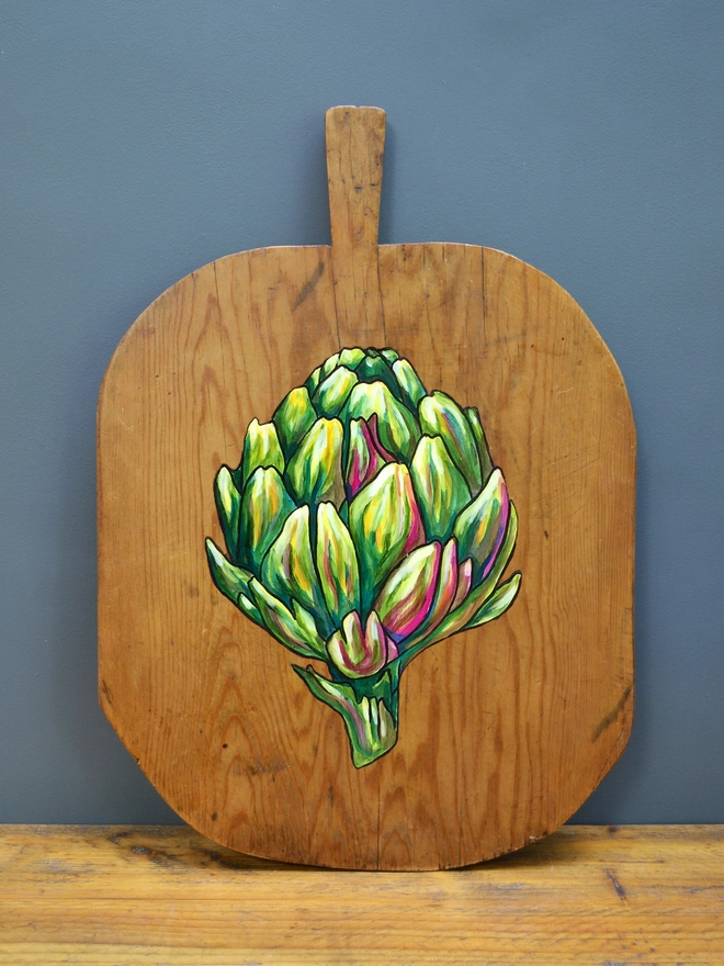 Wooden chopping board with handpainted design of an artichoke standing against a wall