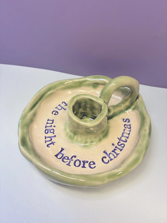 Handmade ceramic Christmas taper candlestick holder with a soft green glazed edge and the words “‘Twas the night before Christmas” in dark blue lettering, arranged around the candle holder opening. Photographed against a pale background.