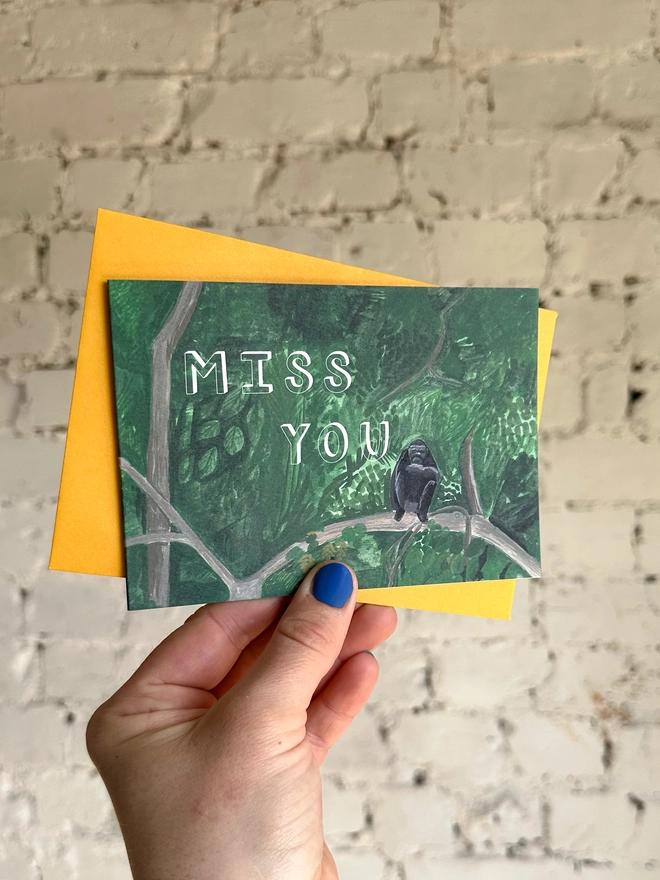 a greetings card featuring an illustration of a chimp sitting in a jungle landscape with the text “miss you” with a deep yellow envelope