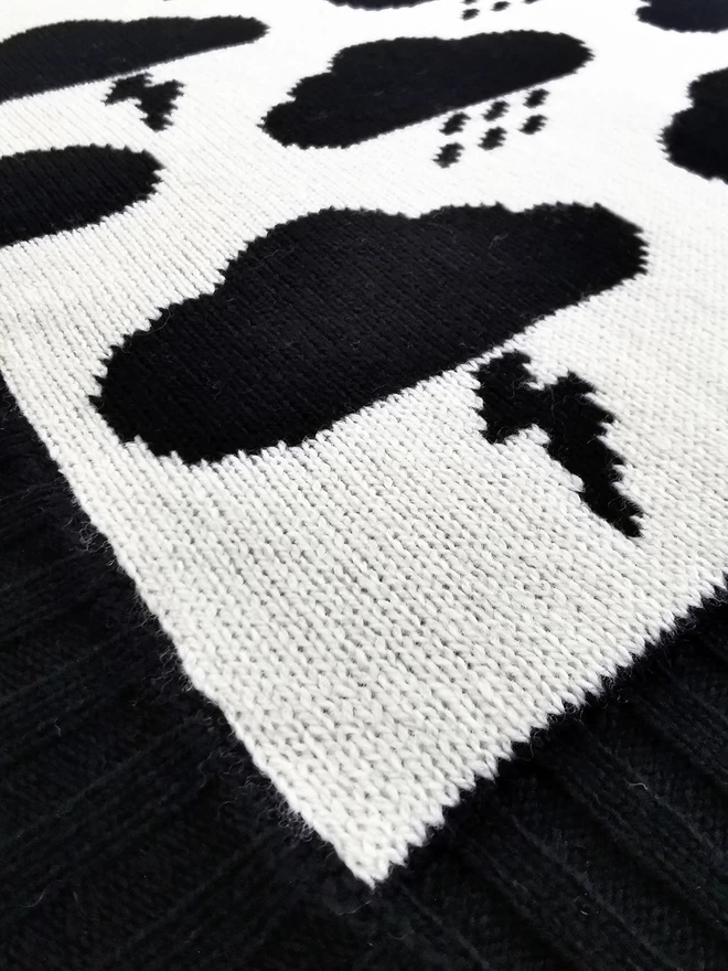 A close up of the corner of a knitted blanket showing the knit stitches, black and white storm cloud design and black ribbed trim.