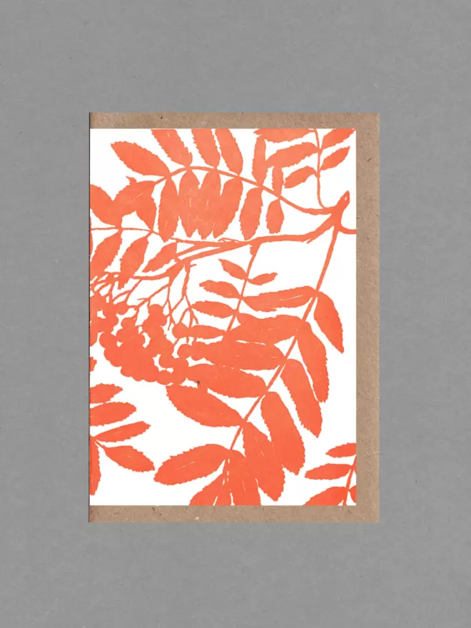 Greetings card with an illustration of a Rowan tree in orange. Behind the card is a brown envelope on a grey backround.