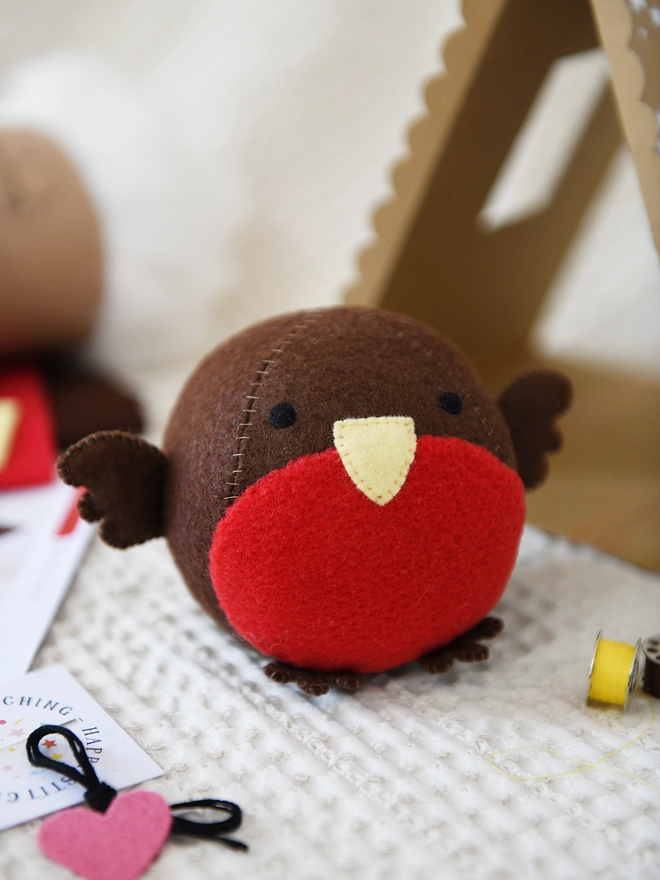 A round robin plush toy stands on an ivory fabric surface with craft kit contents beside it. A cardboard tent house is behind the robin.