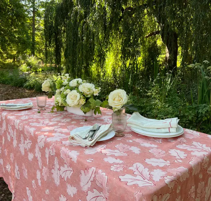 Grace Oak Trees Pink Linen Tablecloth. Light Pink and white tablecloth with oak patterns, styled on an outdoor dining table with plates, glassware, and white floral arrangements.