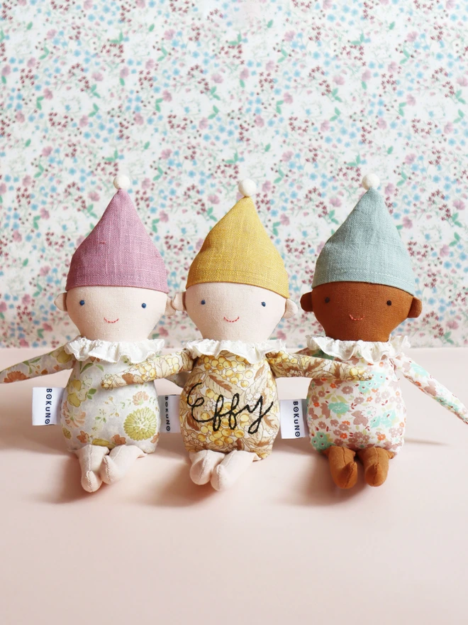 Three handmade Spring Elf Dolls are sitting side by side against a soft pastel background. Each doll has a floral print body, pastel-coloured hat with a pompom, and a ruffled white collar. They feature different skin tones and gentle smiles, radiating charm and warmth.