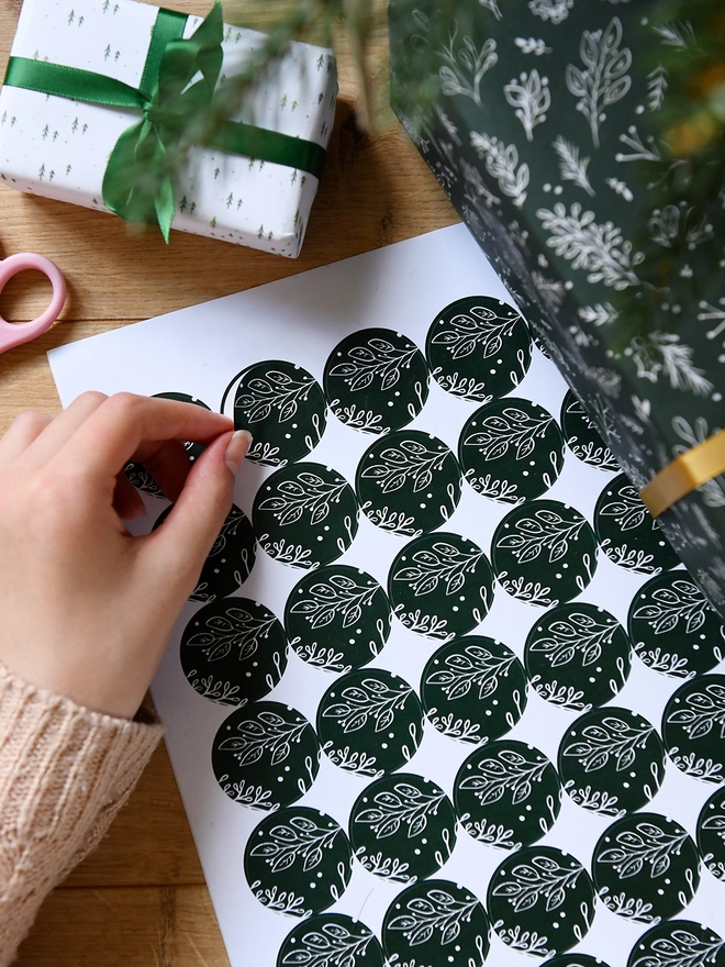 A hand peels a deep green sticker with a Christmas botanical design from a sheet of 35 matching stickers.