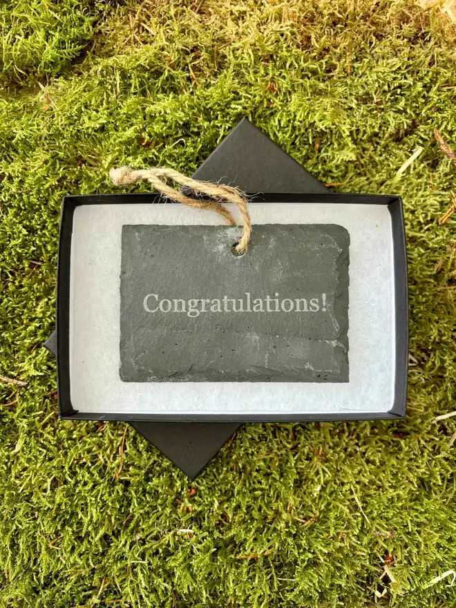 Engraved Congratulations message on a hanging slate sign in a gift box on bed of moss. 