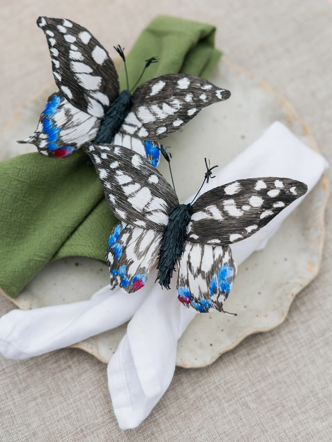 Blue and white patterned handmade butterflies on napkin rings as part of an Easter table, with green and white napkins, on a textured plate and beige tablecloth