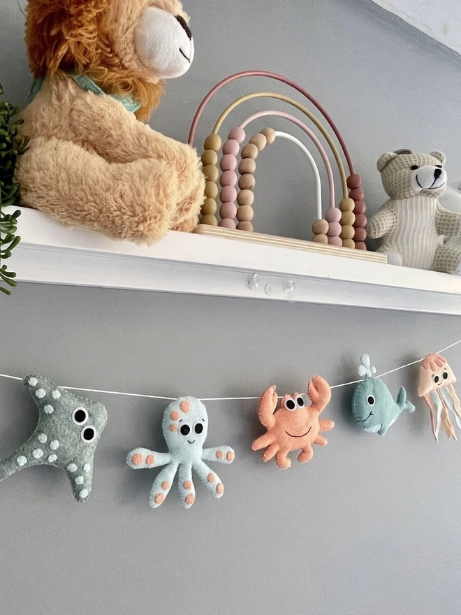 Nursery Under The Sea Garland, a sea themed garland hanging from a white shelf against a grey backdrop in a child's bedroom. 