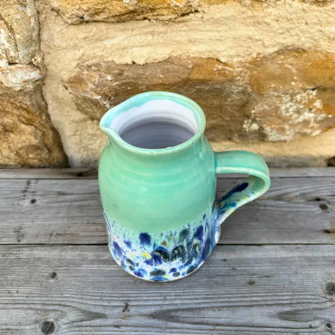 Shoreline Medium Jug, a colourful jug sitting on a wooden surface against a brick wall backdrop. 