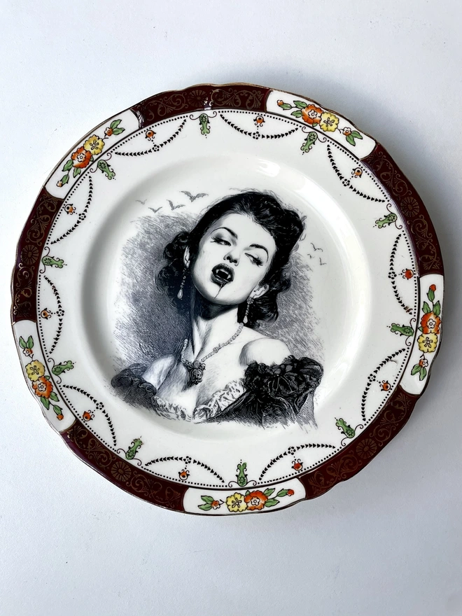 A vampire with bats behind her, the image is victorian in black and white and is printed directly onto an ornate vintage plate