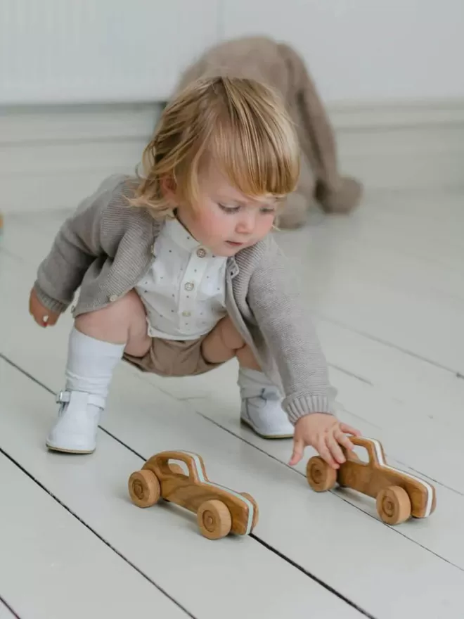 little baby playing with two wooden toy cars in a living room with a wooden floor